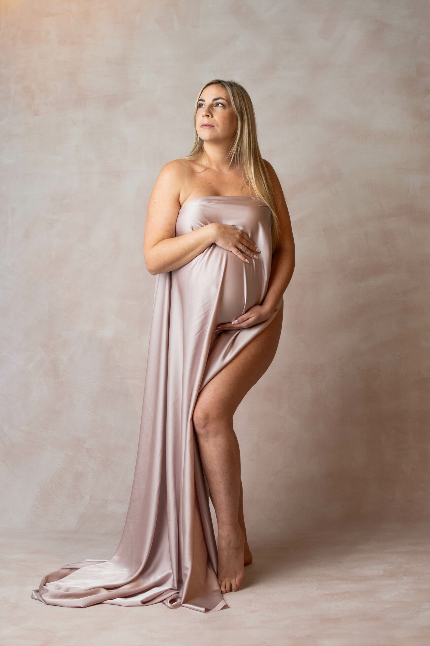 Femme enceinte posant en studio avec un tissu satiné rose poudré sur fond beige texturé.